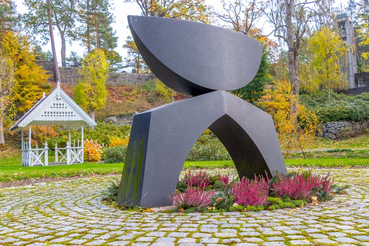Skulptur i anonym minnelund på Arendal kirkegård, med paviljong i bakgrunnen.