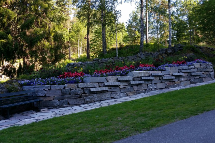 Navneplater på natursteinsmur omgitt av blomster i navnet minnelund på Arendal kirkegård.