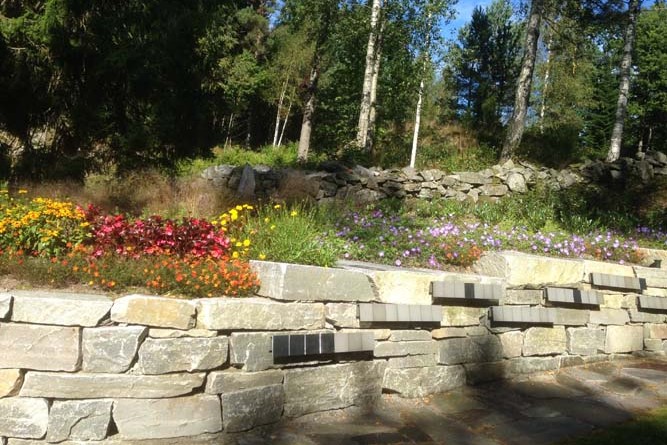 Navnet minnelund på Arendal kirkegård med navneplater, benk og blomster i natursteinsmur langs skogkant.