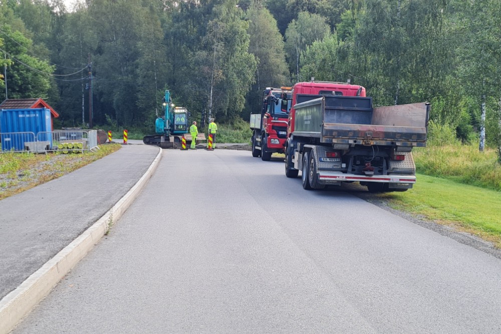 Veiarbeid ved innkjørselen til Arendal kirkegård, med gravemaskin, lastebiler og arbeidere i refleksvester. Kjøretøyene sperrer deler av veien, mens sideområdet og gangfeltet er fritt. Arbeidet kan medføre hindringer for kjøring til parkeringsplassen på dager uten seremonier. Foto.