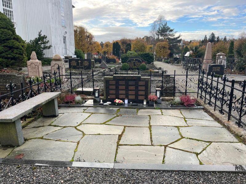 Navnet minnelund på Hisøy kirkegård med navneplater, blomster og benk innenfor smijernsgjerde.